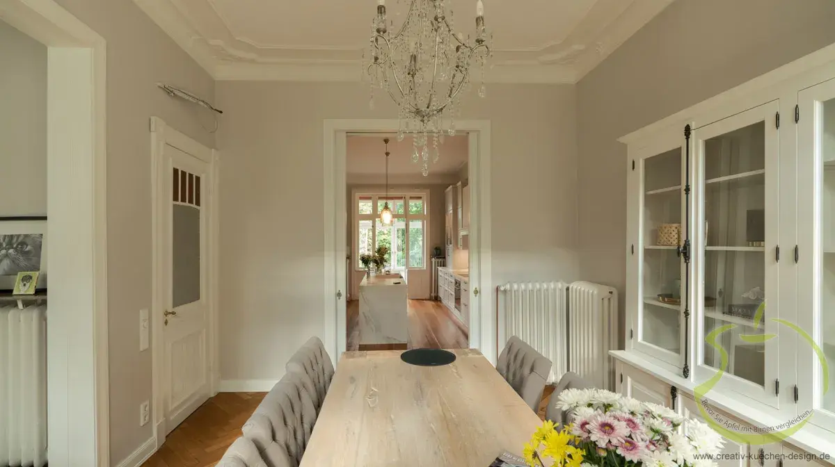 Heller Essbereich mit Holztisch, Blumenstrauß und Kronleuchter, Blick ins Wohnzimmer.