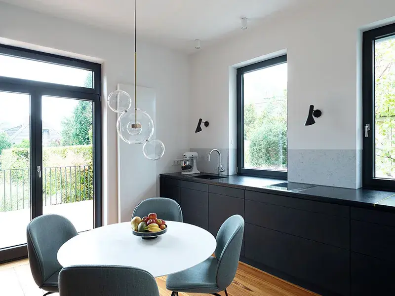 Moderne Küche mit schwarzer Arbeitsplatte, rundem Tisch und Obstschale, große Fenster.