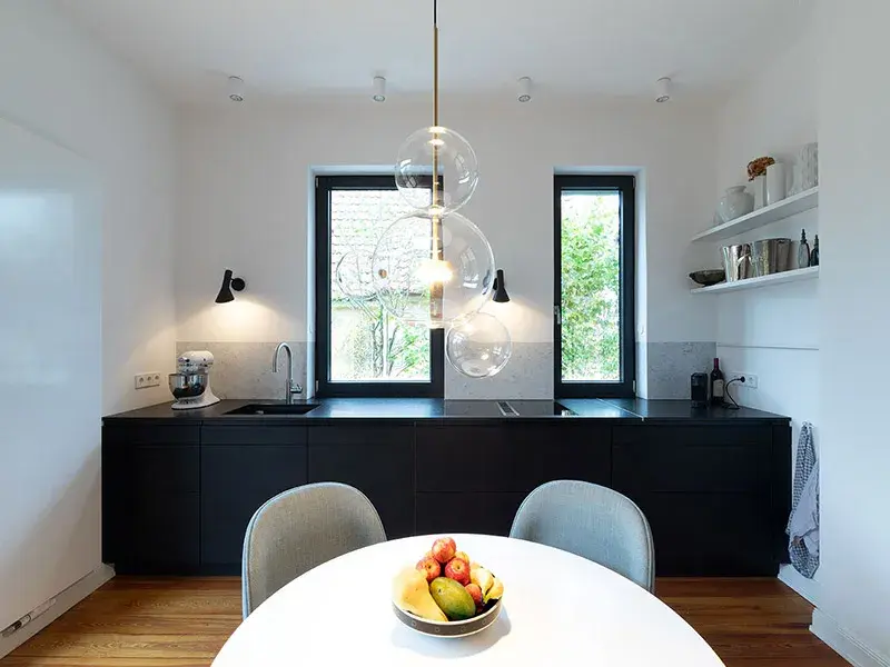 Moderne Küche mit schwarzer Arbeitsplatte, runden Lampen und Obstschale auf weißem Tisch.